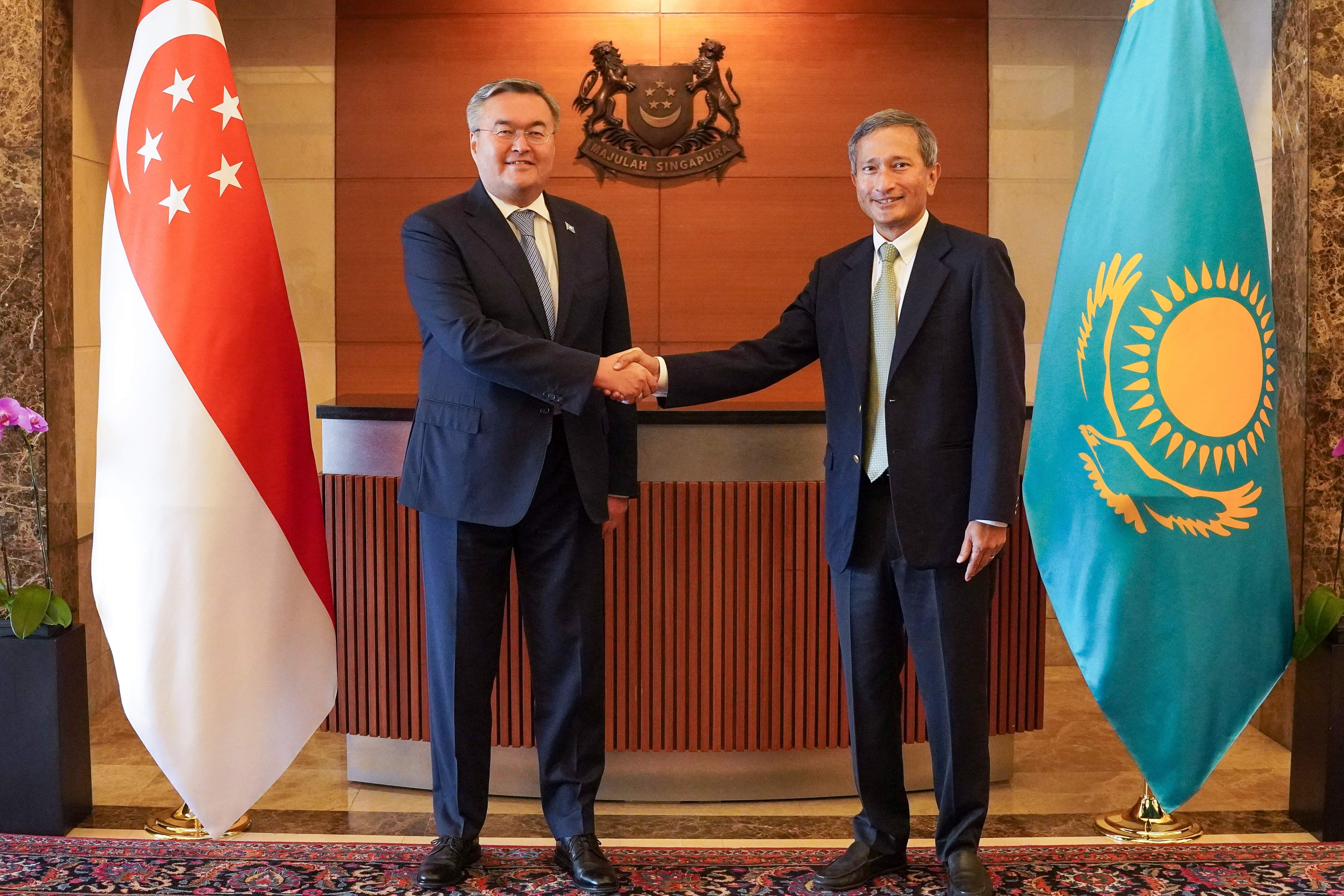 Two men in suits shake hands, standing before the flags of Singapore and Kazakhstan.
